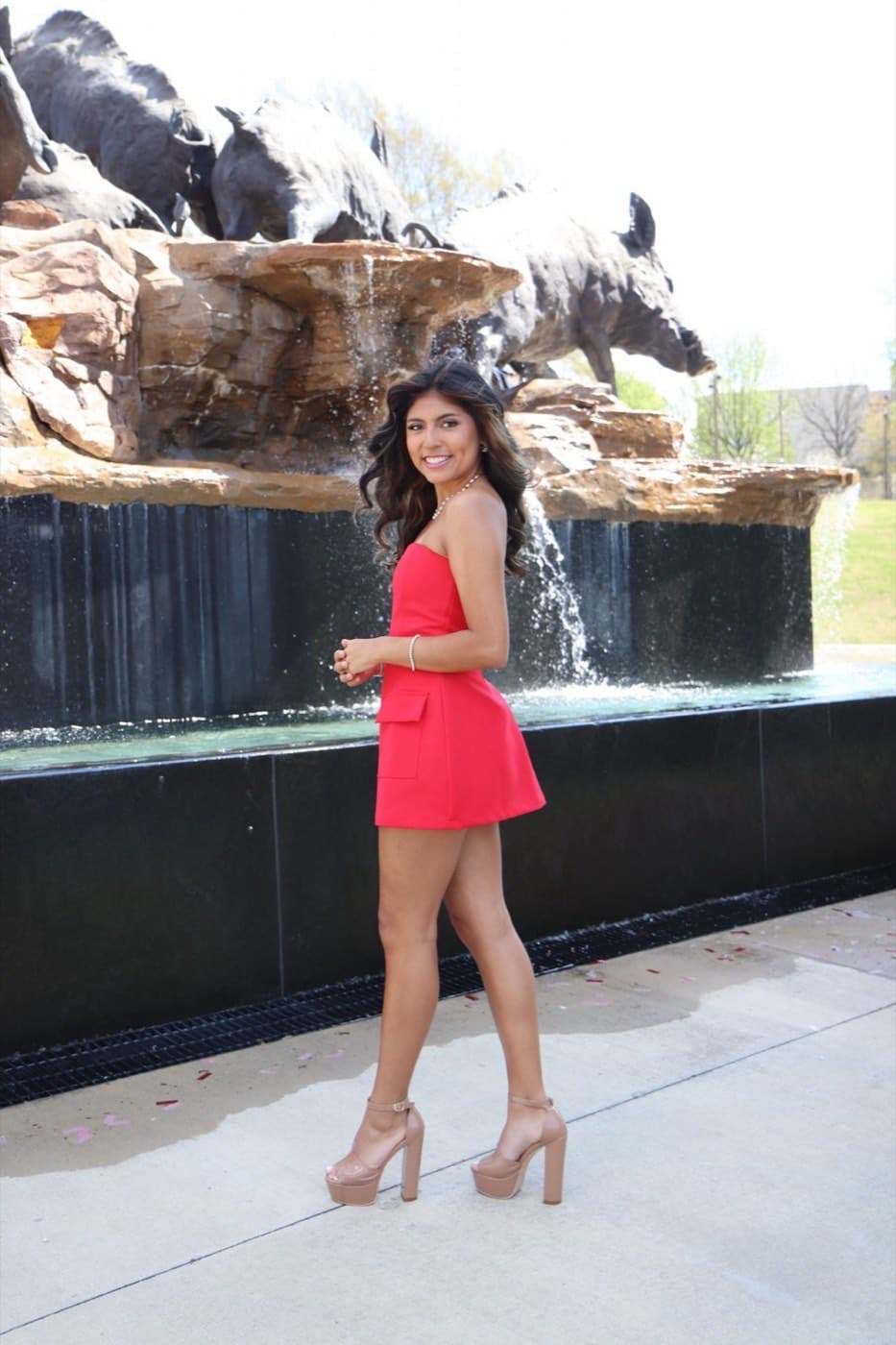 A woman in a red dress and high heels stands smiling near a black stone fountain with statues of wild boars on rocks behind her.